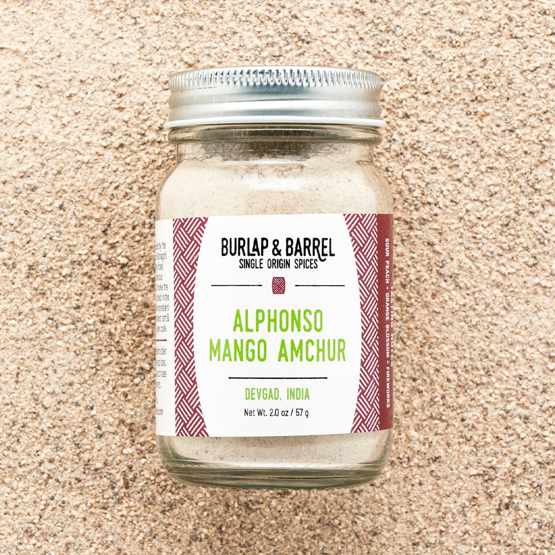 A jar of Alphonso Mango Amchur spice on a background of loose amchur powder with the label 'Burlap & Barrel - Alphonso Mango Amchur
