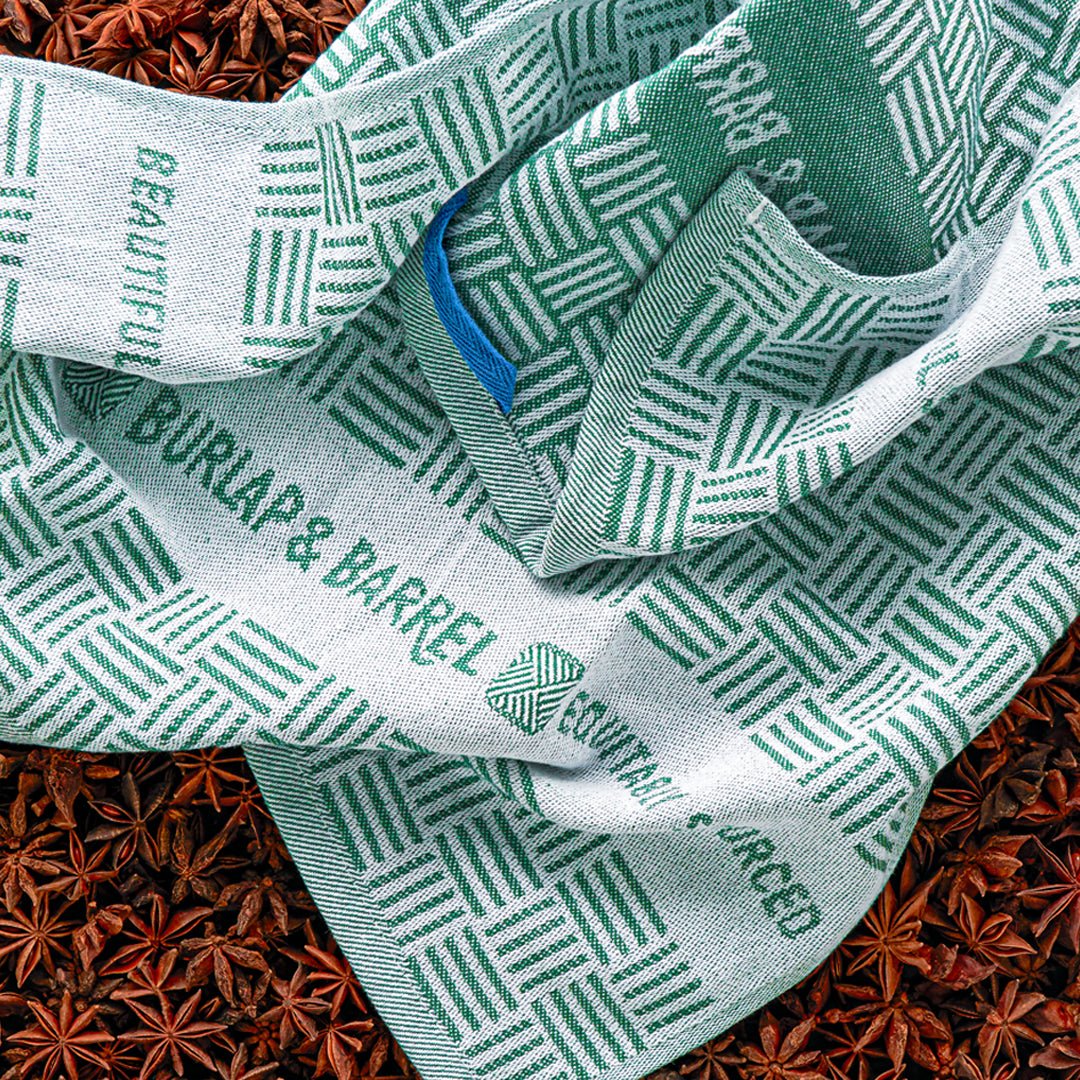 Green and white geometric patterned kitchen towels reading ""BURLAP & BARREL"" arranged on star anise spices.
