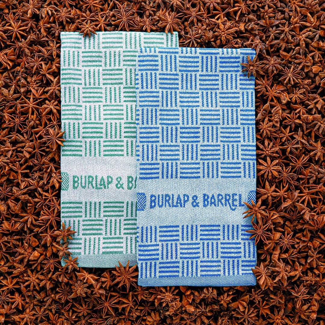 Two folded kitchen towels with geometric stripe patterns in green and blue, branded ""Burlap & Barrel,"" displayed on star anise background.