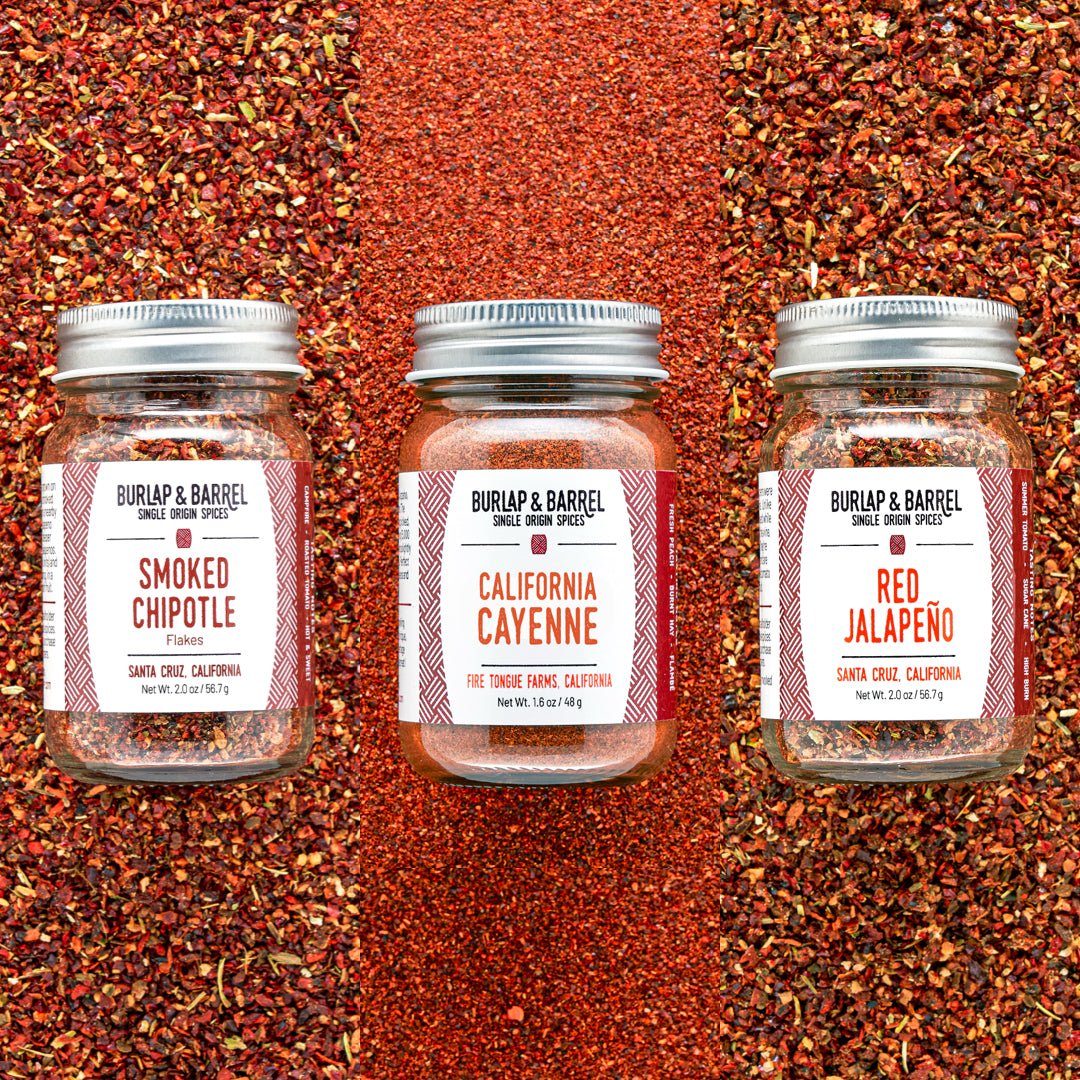 Three glass jars of Burlap & Barrel single-ingredient spices arranged side by side on a background of matching red chili powder. The jars are labeled Smoked Chipotle, California Cayenne, and Red Jalapeño.