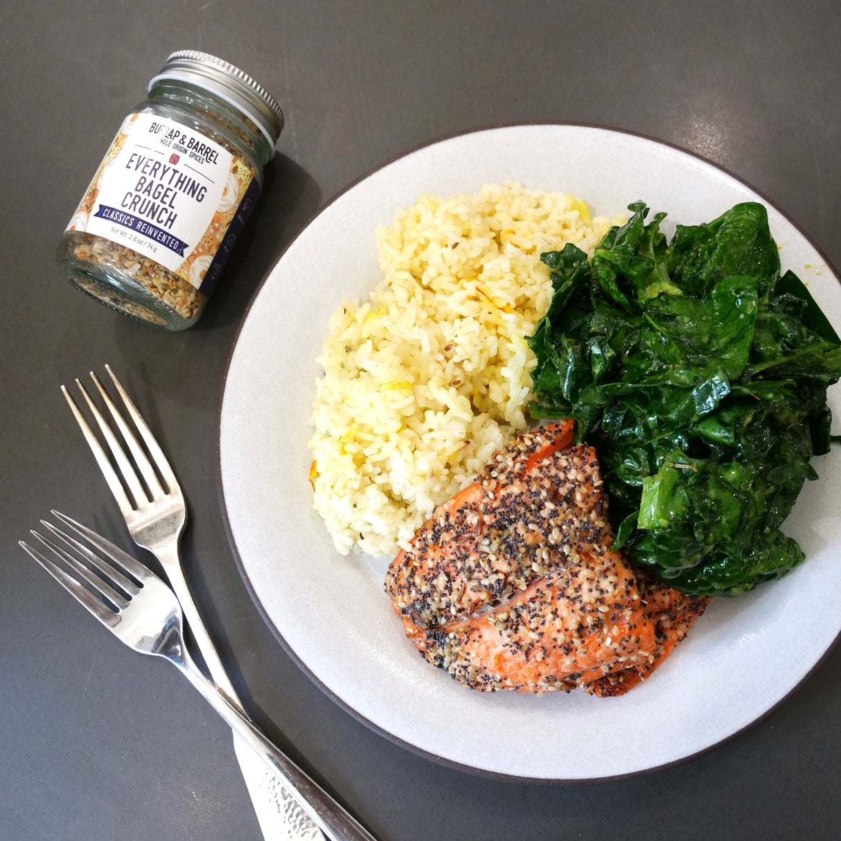 Plate of food with salmon, cauliflower rice, and greens, accompanied by a jar of seasoning on a dark surface.