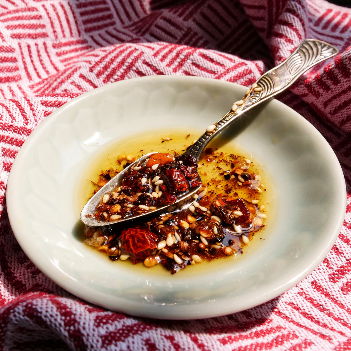 chili oil with dried chili peppers and garlic in a white bowl with a spoon on a red and white patterned cloth.