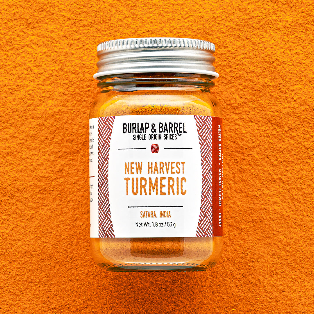 Glass jar of New Harvest Turmeric from Burlap & Barrel with a metal lid on a background of loose turmeric powder.