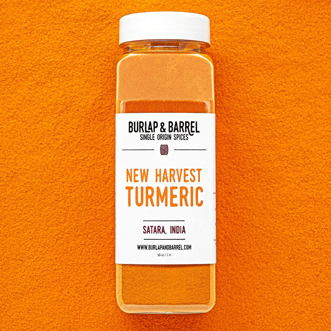 Plastic container of New Harvest Turmeric from Burlap & Barrel with a white lid on a background of loose turmeric powder.