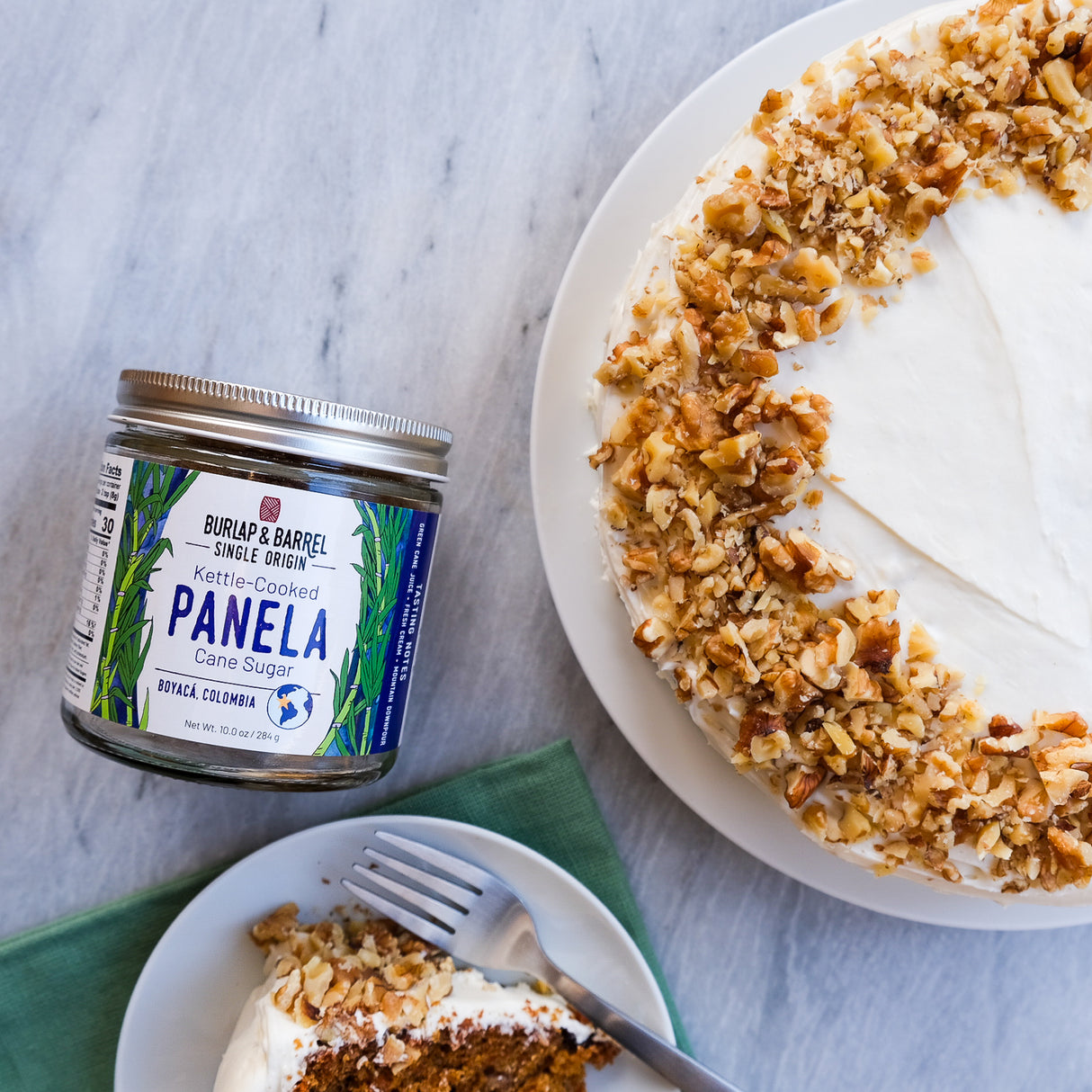 Jar of Burlap & Barrel Panela sugar next to a cake with a slice taken out on a marble surface.