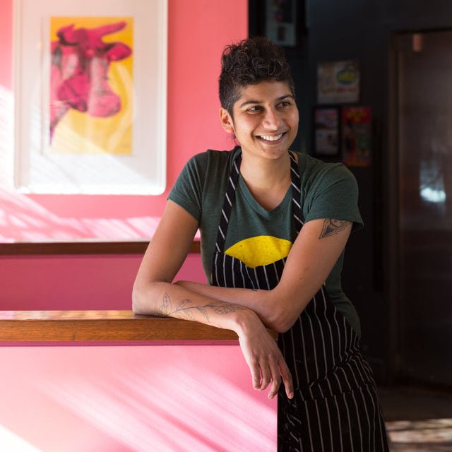 Preeti Mistry smiling at counter in vibrant pink restaurant with colorful artwork on wall