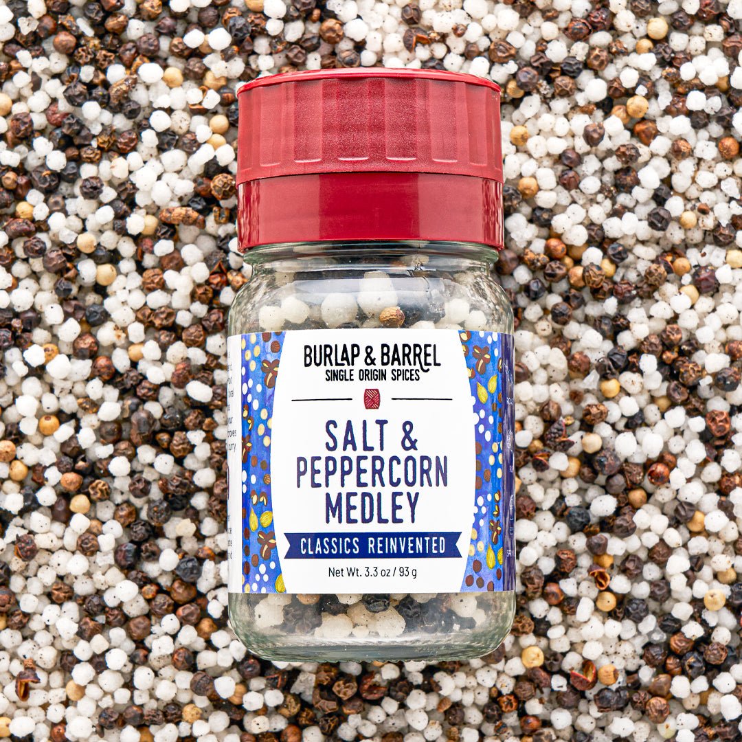 A bottle of Burlap & Barrel Salt & Peppercorn Medley spices, featuring a red cap and a white label with blue patterns, set against a background of mixed whole peppercorns.