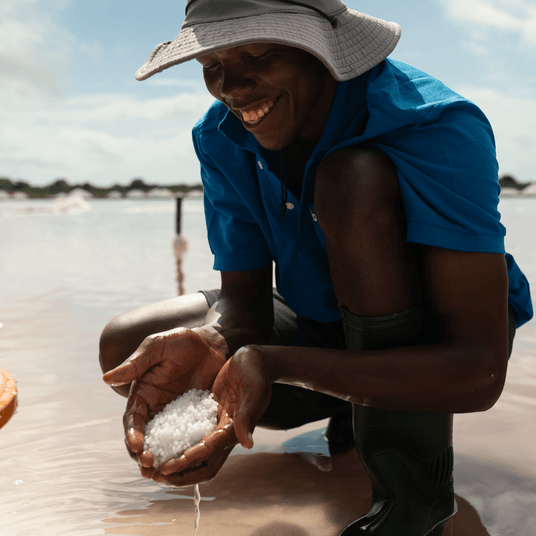 Partner farmer smiling in a blue shirt and tan hat, holding two handfuls of hand harvested pearl salt in Tanzania.