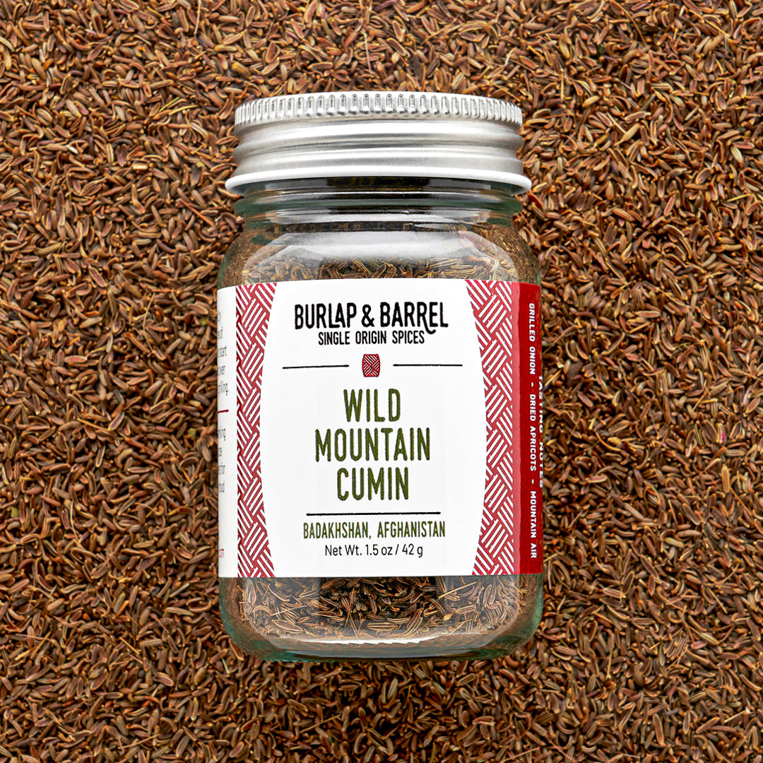 Glass jar of wild mountain cumin from Burlap & Barrel with a metal lid on a background of loose cumin seeds.