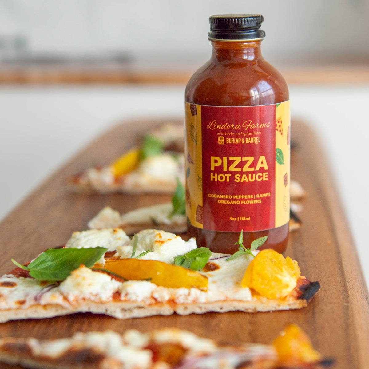 Lindera Farms Pizza Hot Sauce bottle next to fresh pizza with mozzarella and yellow peppers on wooden board.