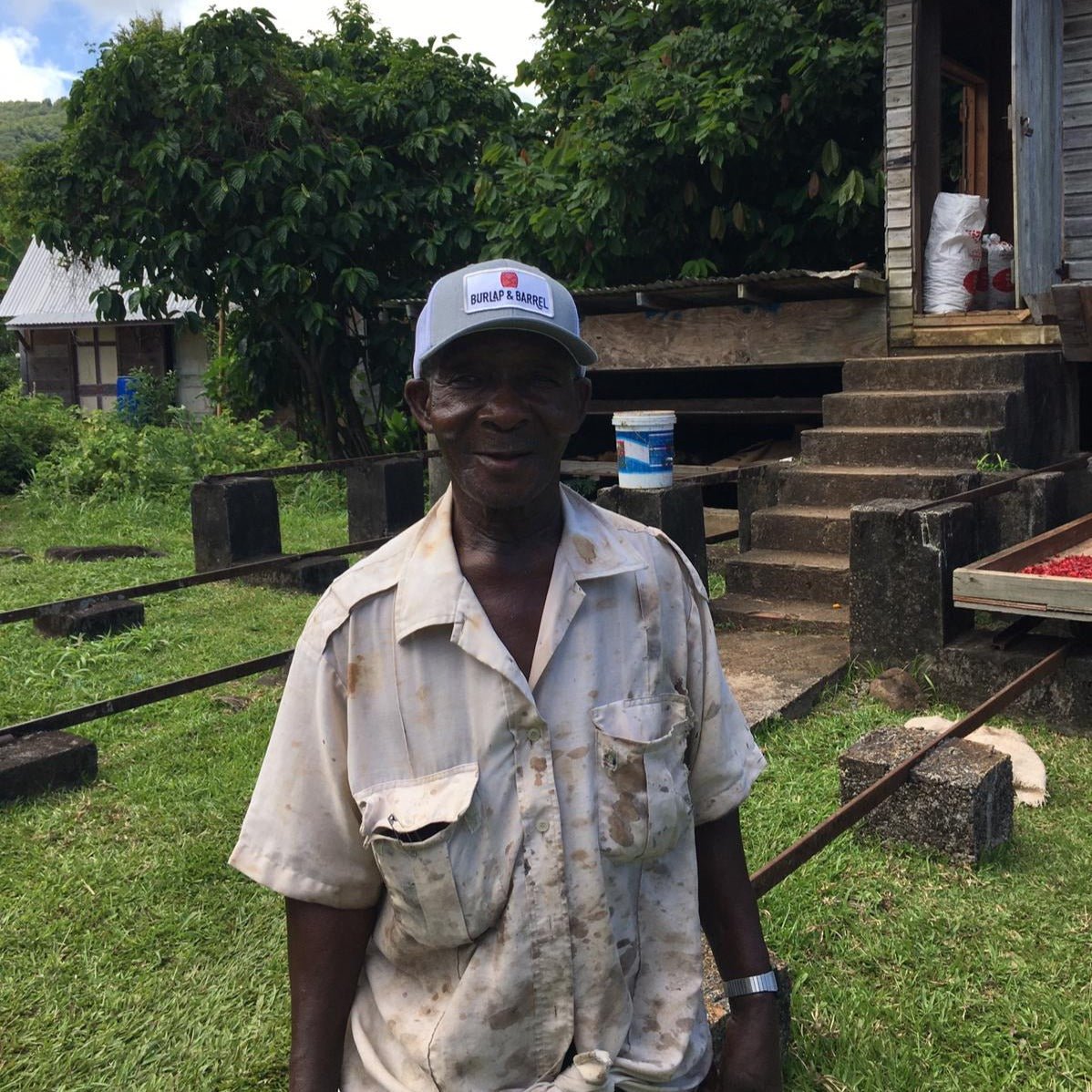 A smiling man wearing a ""Burlap & Barrel"" cap and light work shirt stands in front of a rural property with lush vegetation.