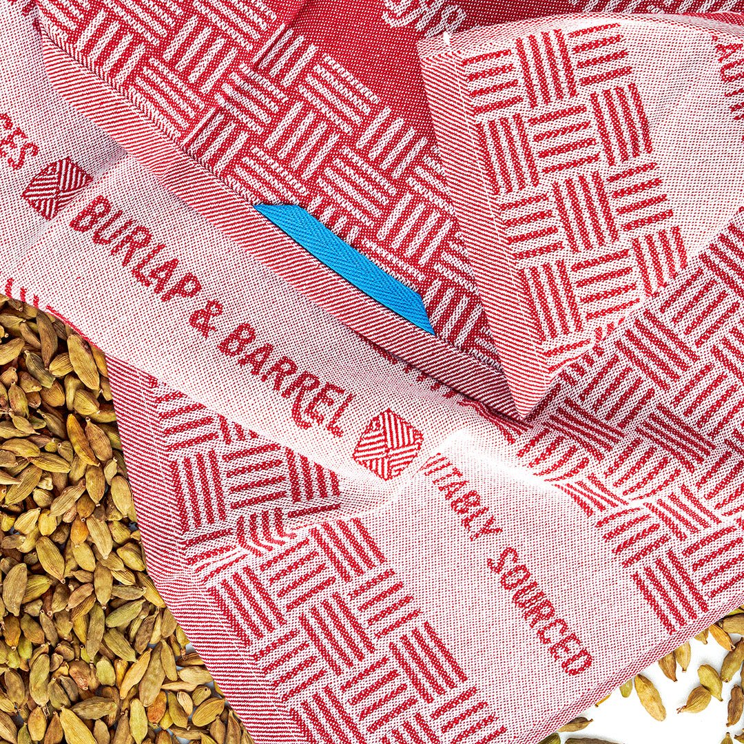 Red and white patterned kitchen towel with ""BURLAP & BARREL"" text, blue feather, and scattered cardamom pods.