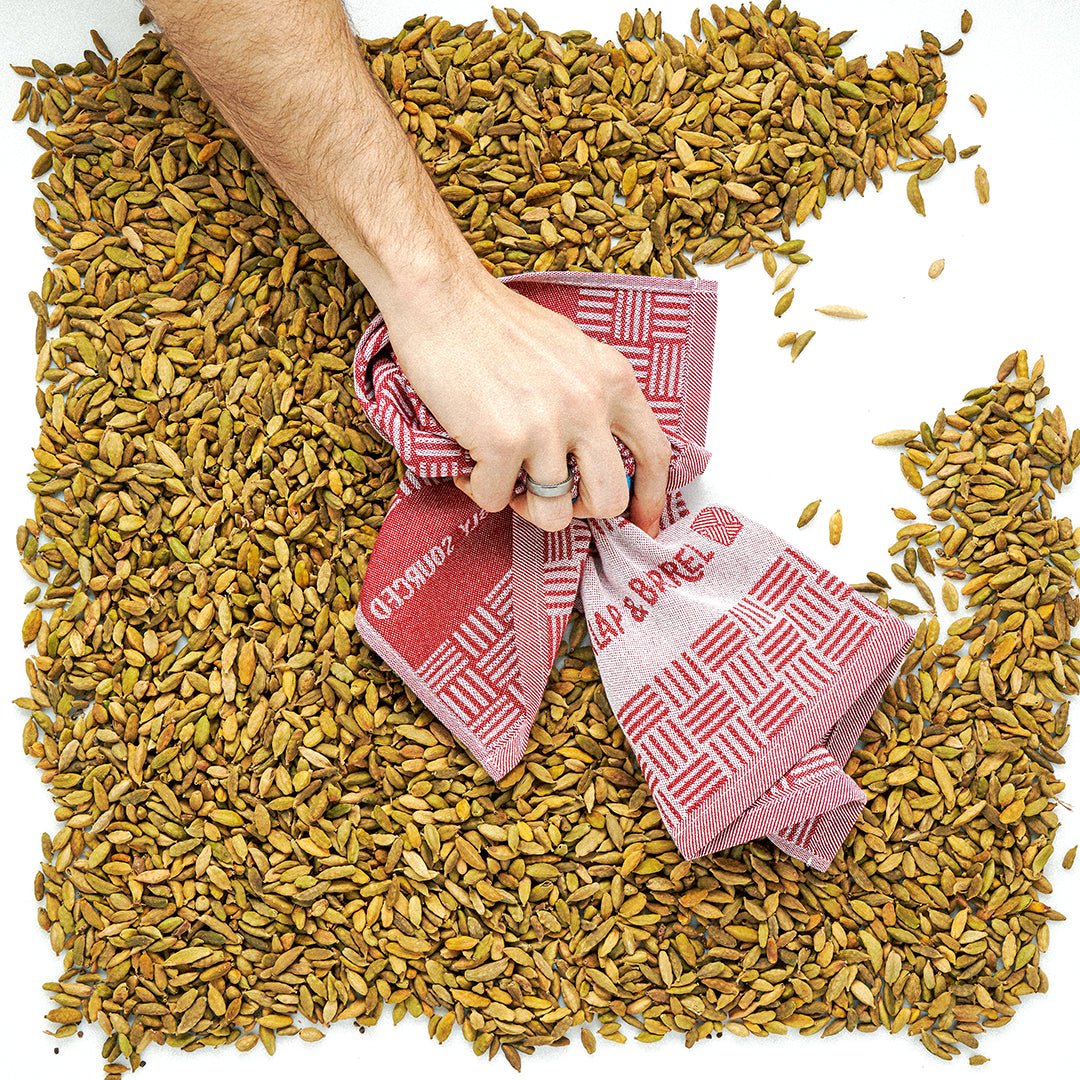 A hand holding red and white patterned kitchen towels wiping a pile of cardamom pods.
