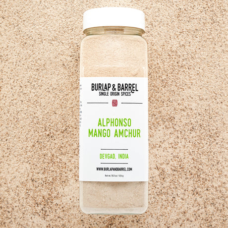 A jar of Alphonso Mango Amchur spice on a background of loose amchur with the label 'BURLAP & BARREL' and 'Alphonso Mango Amchur