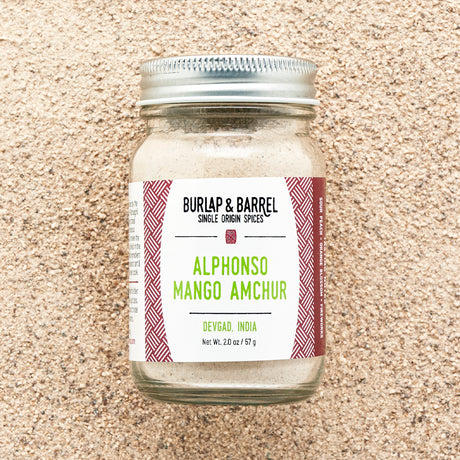 A jar of Alphonso Mango Amchur spice on a background  of loose amchur powder with the label 'Burlap & Barrel - Alphonso Mango Amchur
