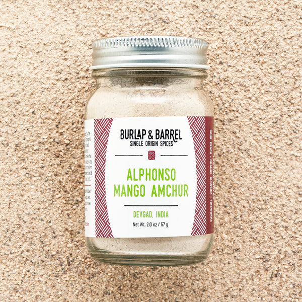 A jar of Alphonso Mango Amchur spice on a background  of loose amchur powder with the label 'Burlap & Barrel - Alphonso Mango Amchur