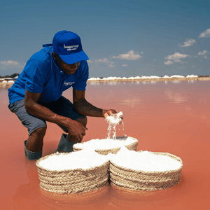 Partner farmer in a blue at and shirt standing in water and hand-harvesting salt in Tanzania. There are three baskets full of salt.