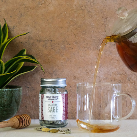 Tea being poured into a glass mug with a jar of 'Anatolian Sage' tea leaves and a honey dipper on a wooden surface.