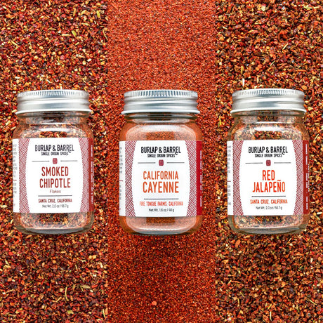Three glass jars of Burlap & Barrel single-ingredient spices arranged side by side on a background of matching red chili powder. The jars are labeled Smoked Chipotle, California Cayenne, and Red Jalapeño.