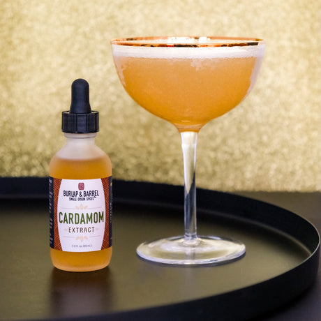 Bottle of Burlap & Barrel Cardamom Extract next to a cocktail glass with an orange drink on a black tray.