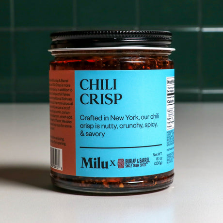 A jar of Chili Crisp with a label indicating it is crafted in New York, by Milu. The jar is placed against a background of green tiles.