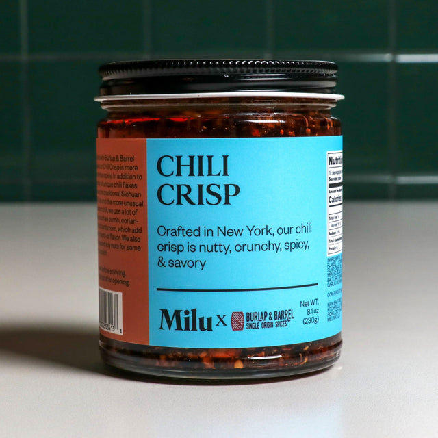 A jar of Chili Crisp with a label indicating it is crafted in New York, by Milu. The jar is placed against a background of green tiles.