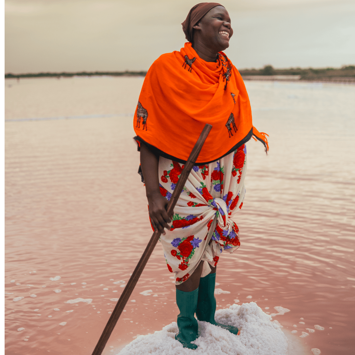 Partner farmer with an orange shawl smiling atop of a pile of salt. Surrounding her is water and she is holding a long stick.