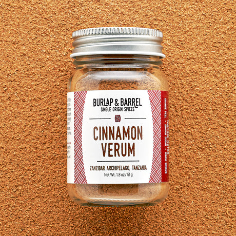 A glass jar of Cinnamon Verum spice with a label that reads 'Cinnamon Verum' and 'Zanzibar Archipelago, Tanzania'. The jar is placed against a textured background.