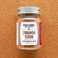 A glass jar of Cinnamon Verum spice with a label that reads 'Cinnamon Verum' and 'Zanzibar Archipelago, Tanzania'. The jar is placed against a textured background.