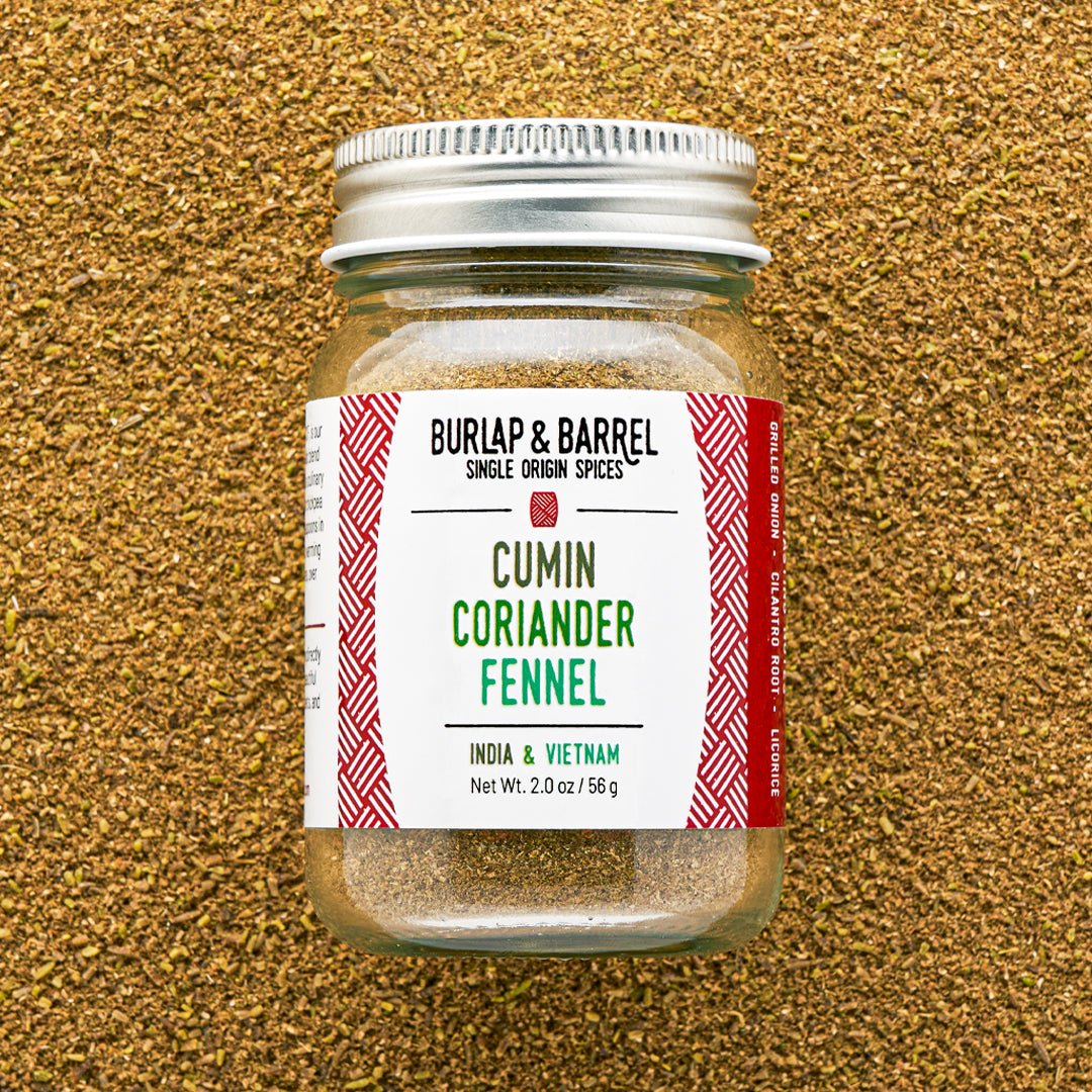 A glass jar of Burlap & Barrel Cumin Coriander Fennel spice mix, featuring a white label with red patterns and a silver metal lid, set against a textured background of loose ground coriander seeds.