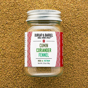 A glass jar of Burlap & Barrel Cumin Coriander Fennel spice mix, featuring a white label with red patterns and a silver metal lid, set against a textured background of loose ground coriander seeds.