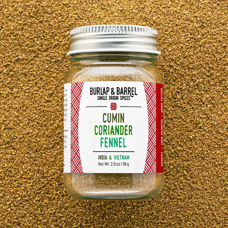 A glass jar of Burlap & Barrel Cumin Coriander Fennel spice mix, featuring a white label with red patterns and a silver metal lid, set against a textured background of loose ground coriander seeds.