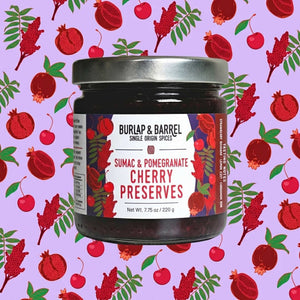 A jar of Burlap & Barrel Sumac & Pomegranate Cherry Preserves centered on a light purple background patterned with red cherries and pomegranates. The jar has a silver metal lid and a white label with red and black text.
