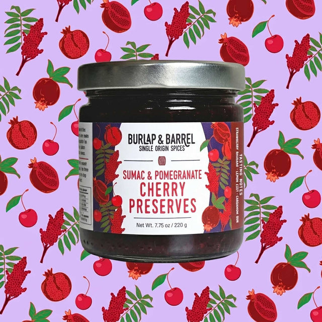 A jar of Burlap & Barrel Sumac & Pomegranate Cherry Preserves centered on a light purple background patterned with red cherries and pomegranates. The jar has a silver metal lid and a white label with red and black text.