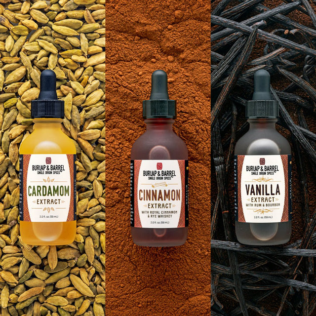 Three bottles of Burlap & Barrel spice extract syrup shown side by side: Cardamom Extract in a yellow bottle over cardamom seeds, Cinnamon Extract in a brown bottle over cinnamon powder, and Vanilla Extract in a dark bottle over vanilla beans.
