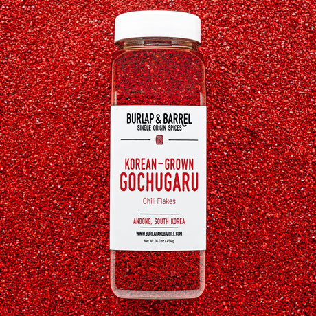 Container of Burlap & Barrel Korean-Grown Gochugaru Chili Flakes on a red background of loose chili flakes