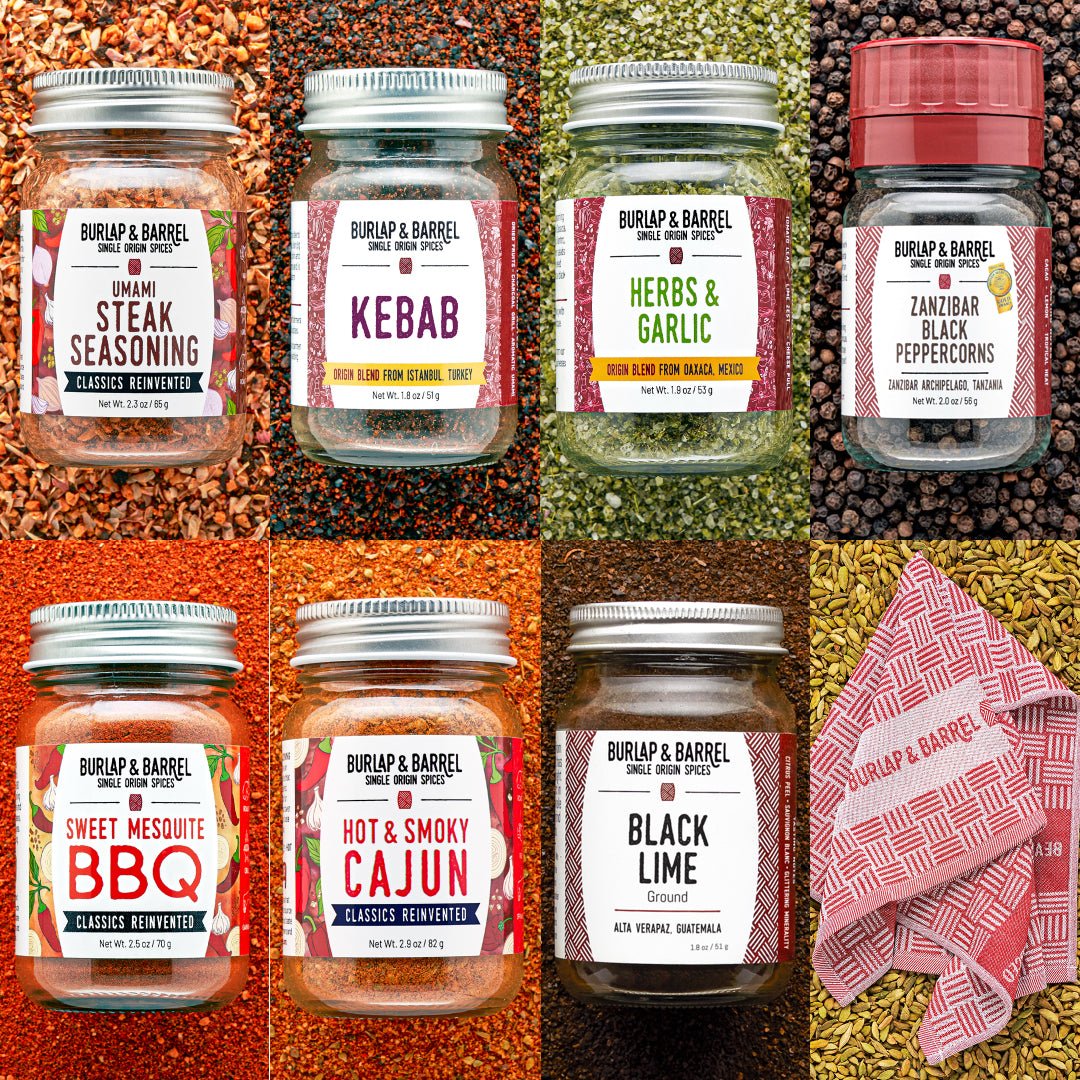 Six glass spice jars from Burlap & Barrel arranged in a 2x3 grid over colorful whole spices and a pink patterned cloth. The jars include Steak Seasoning, Kebab, Herbs & Garlic, Zanzibar Black Peppercorns, Sweet Mesquite BBQ, and Hot & Smoky Cajun.