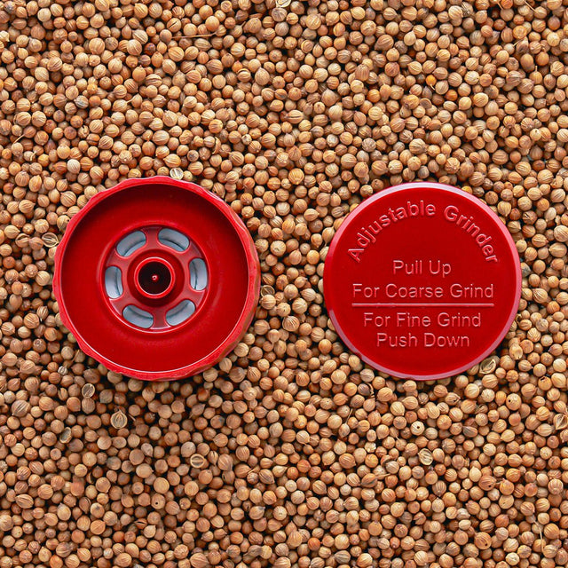 A red adjustable grinder top placed on a background of seeds, with instructions 'Pull Up For Coarse Grind' and 'Push Down For Fine Grind' visible.