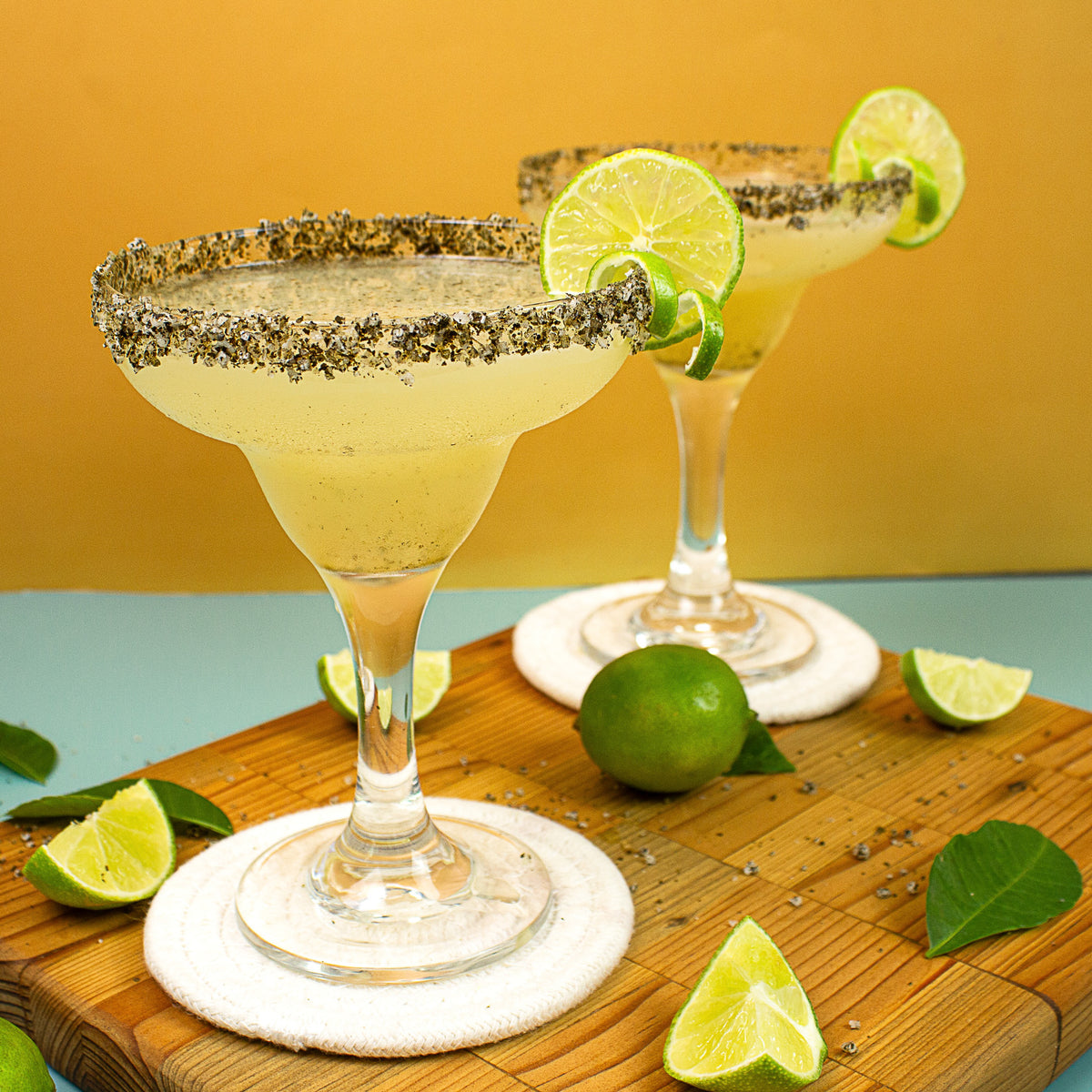 Two margarita glasses with lime wedges on a wooden cutting board against a yellow background