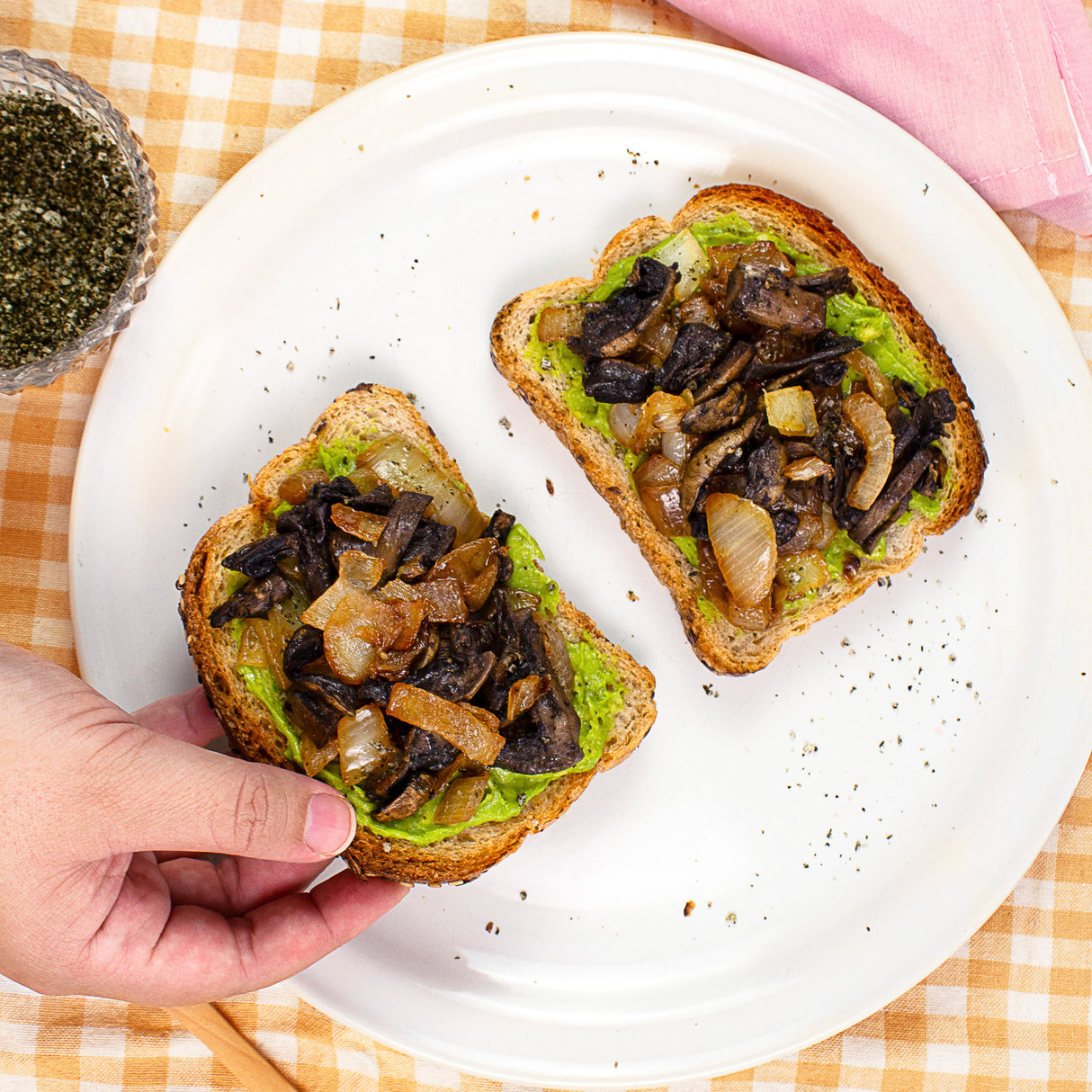 Two slices of toast with avocado and mushrooms on a white plate, with a hand holding one slice.