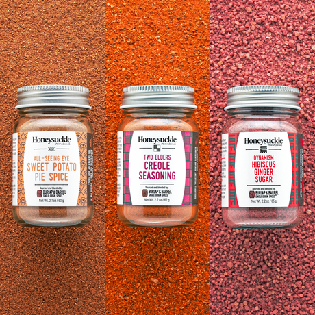 Three glass jars of Honeysuckle brand spice blends, arranged on a textured red background. The jars are labeled as 'SWEET POTATO PIE SPICE,' 'CREOLE SEASONING,' and 'HIBISCUS GINGER SUGAR.