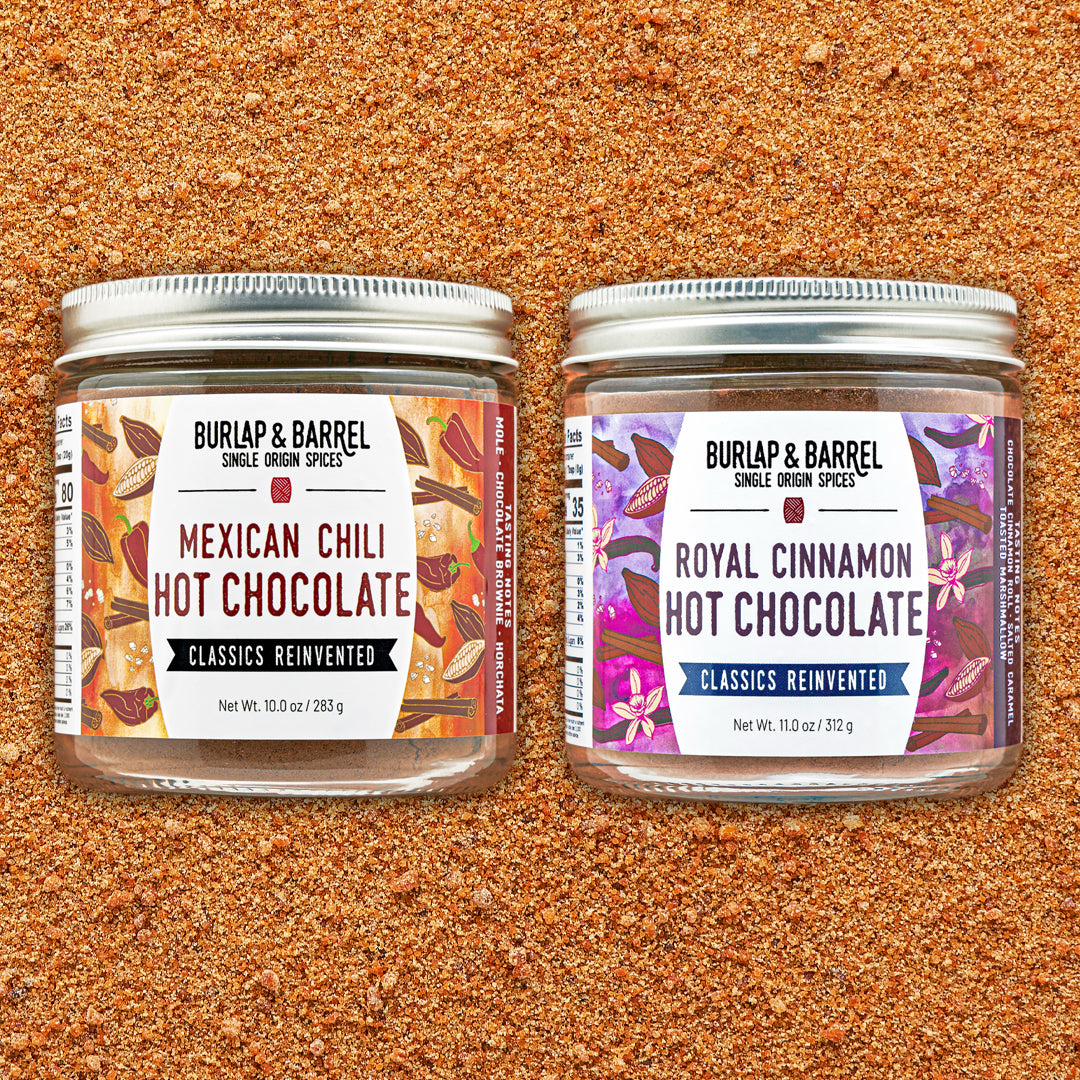 Two glass spice jars with silver lids on a textured brown ground, labeled Burlap & Barrel Mexican Chili Hot Chocolate and Royal Cinnamon Hot Chocolate.