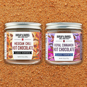 Two glass spice jars with silver lids on a textured brown ground, labeled Burlap & Barrel Mexican Chili Hot Chocolate and Royal Cinnamon Hot Chocolate.