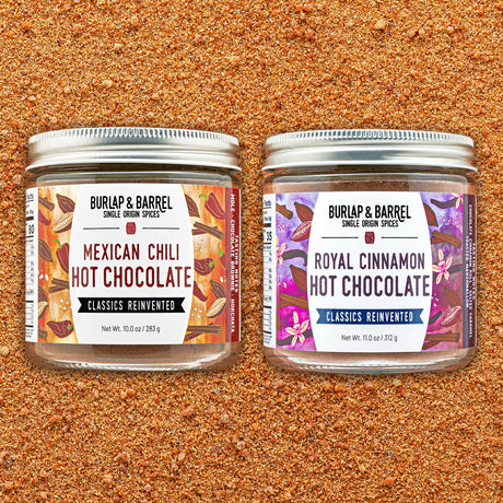 Two glass spice jars with silver lids on a textured brown ground, labeled Burlap & Barrel Mexican Chili Hot Chocolate and Royal Cinnamon Hot Chocolate.