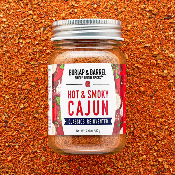 A glass jar of Burlap & Barrel Hot & Smoky Cajun seasoning, featuring a white label with red and blue text, set against a background of loose red-orange ground spices.