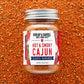 A glass jar of Burlap & Barrel Hot & Smoky Cajun seasoning, featuring a white label with red and blue text, set against a background of loose red-orange ground spices.