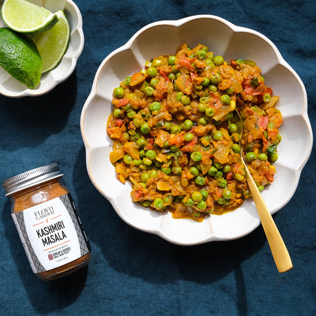 A white bowl with scalloped edges with a golden spoon sitting in a serving of peas and onion with a jar of kashmiri masala next to it.