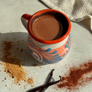 A hand-painted ceramic mug filled with a brown beverage, styled on a light surface with vanilla beans, cinnamon powder, and a textured cloth in the background.
