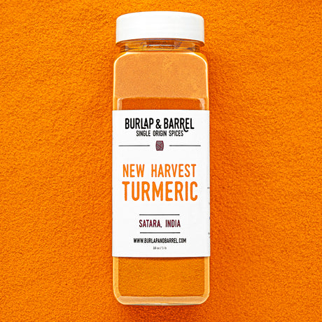 Plastic container of New Harvest Turmeric from Burlap & Barrel with a white lid on a background of loose turmeric powder.