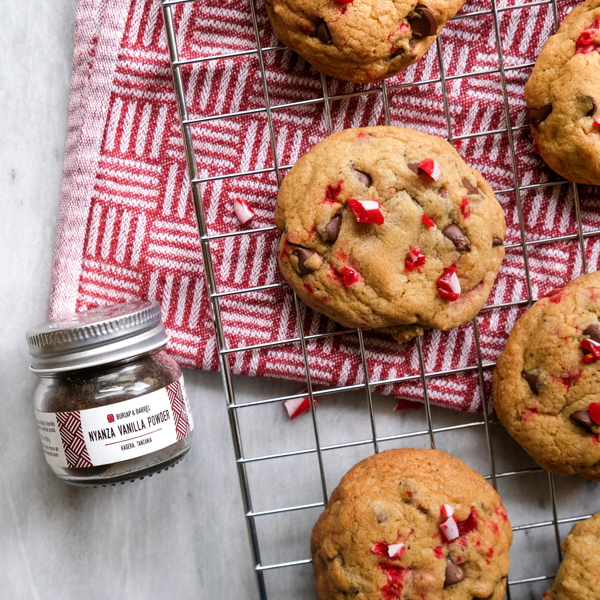 Chocolate chip cookies with red and white candy pieces cooling on a wire rack next to Nyanza Vanilla Powder.